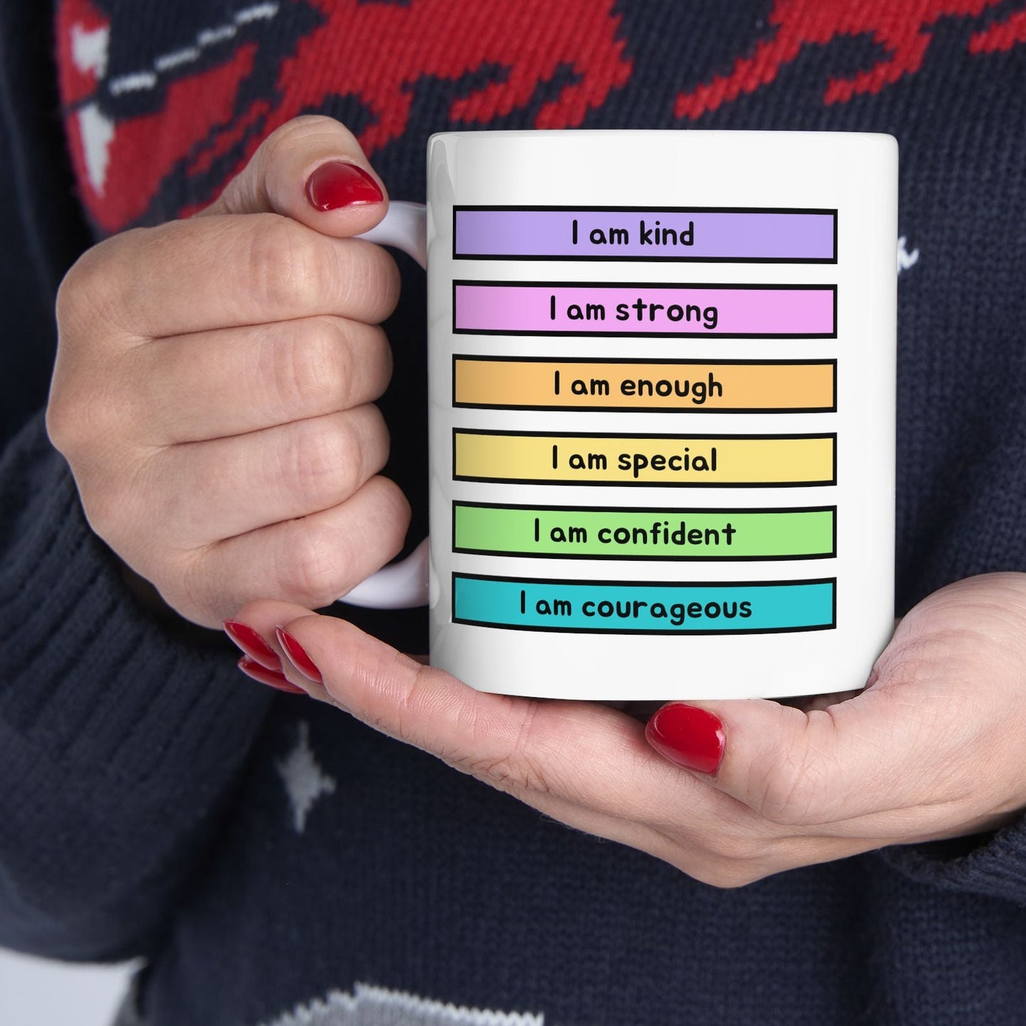 A person holding a white ceramic mug with colorful text blocks reading self-affirmations like 'I am kind', 'I am strong', 'I am enough', 'I am special', 'I am confident', and 'I am courageous'. The person is wearing a dark navy knit sweater with a red and gray holiday pattern.