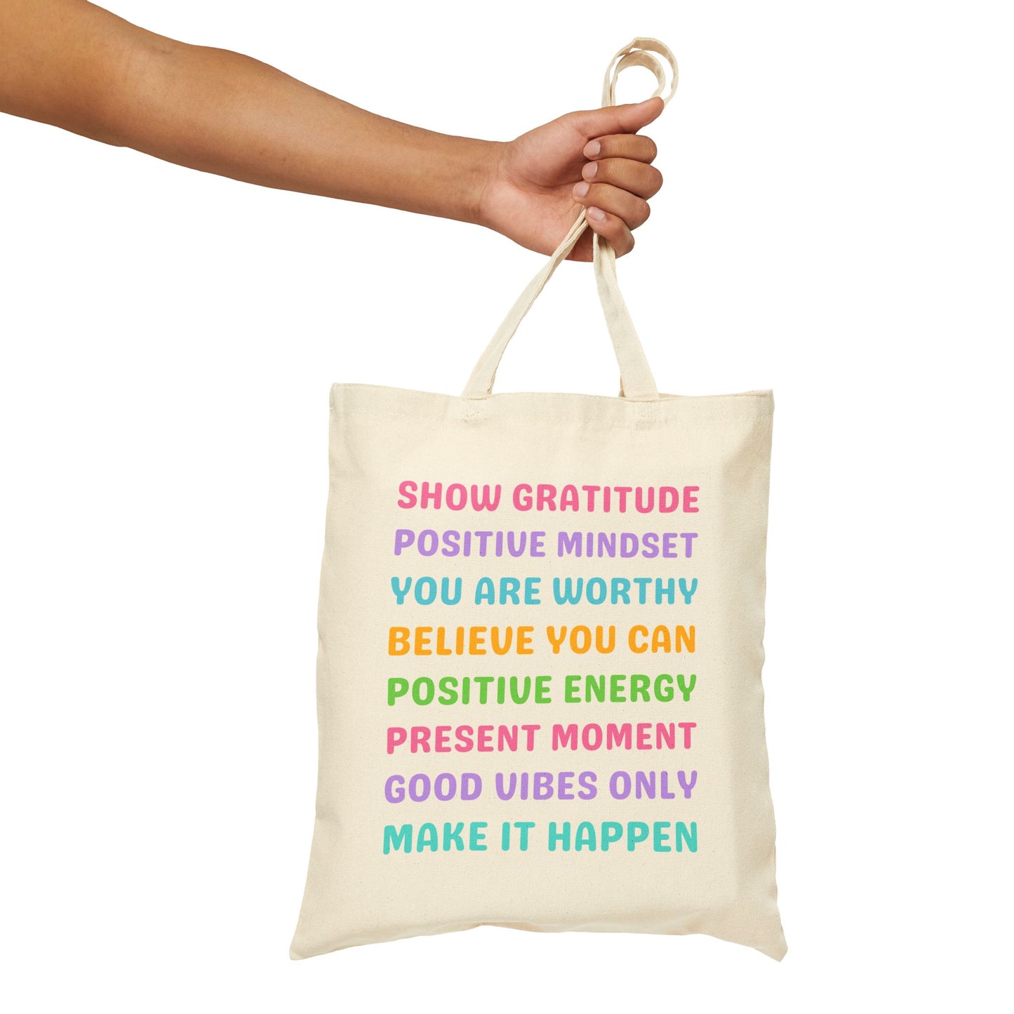 An off-white canvas tote bag with colorful motivational text, held by a person's arm against a plain white background.