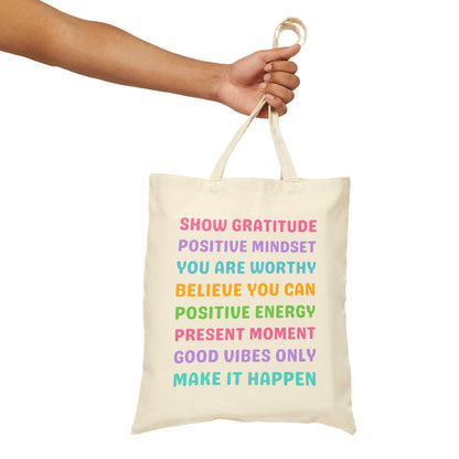 An off-white canvas tote bag with colorful motivational text, held by a person's arm against a plain white background.