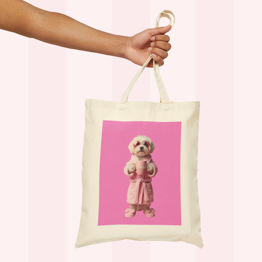An off-white canvas tote bag with a pink square graphic print of a small dog in a pink robe, held by a person's hand against a pale pink striped background.