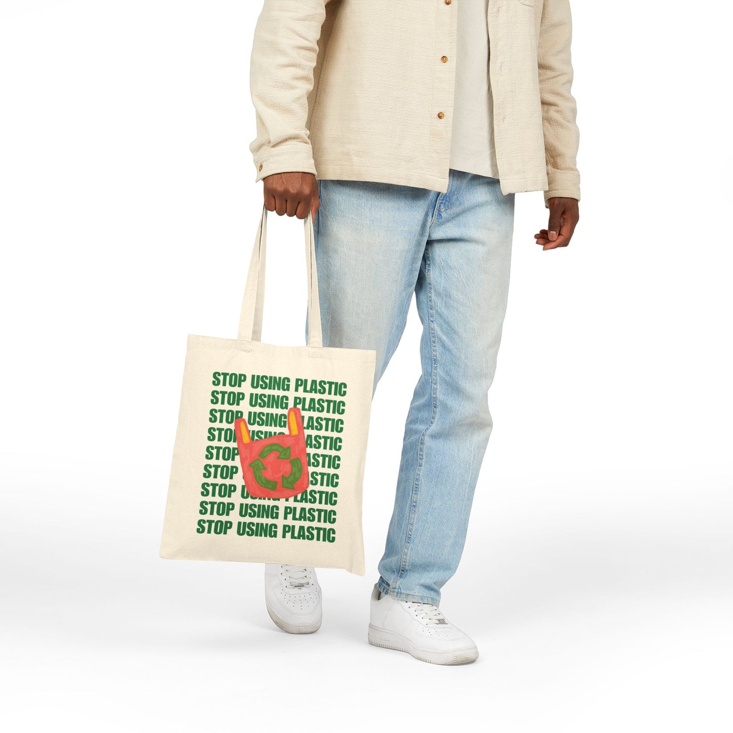 A person wearing a beige button-up shirt, light blue jeans, and white sneakers holds a cream canvas tote bag with green 'STOP USING PLASTIC' text and a red plastic bag icon. The photo is taken against a clean white background.