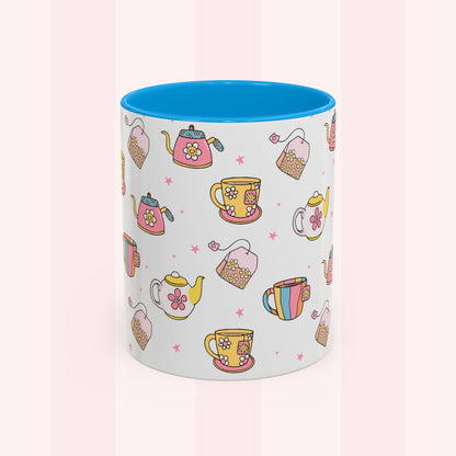 A white ceramic mug with a bright blue interior, decorated with a repeating pattern of colorful teapots, teacups, and tea bags on a soft pink striped background.