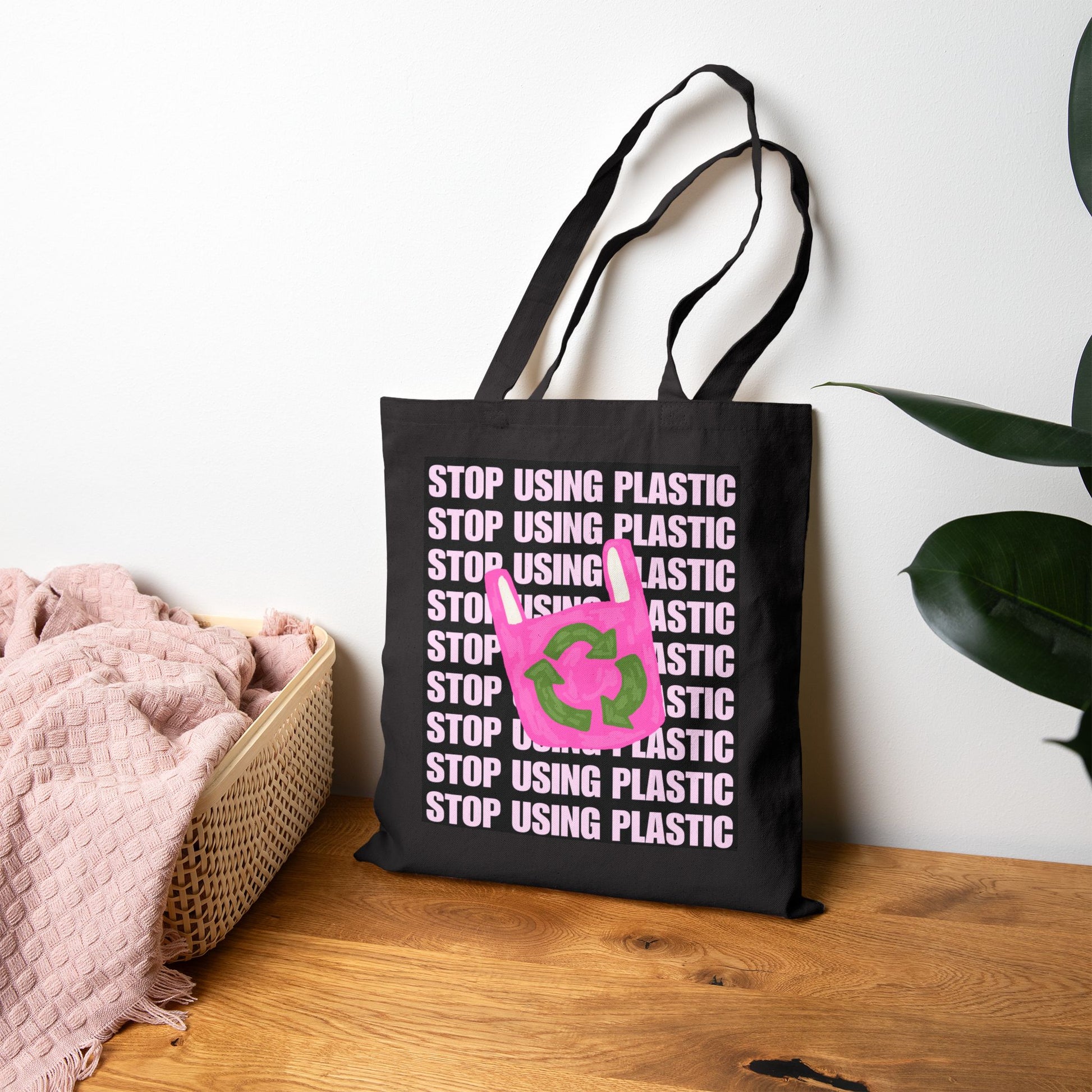 A black canvas tote bag with repeated 'STOP USING PLASTIC' text in white and a pink recycling symbol with green leaves, standing on a wooden surface against a white wall with a basket and plant nearby.