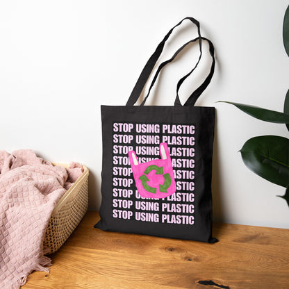 A black canvas tote bag with repeated 'STOP USING PLASTIC' text in white and a pink recycling symbol with green leaves, standing on a wooden surface against a white wall with a basket and plant nearby.