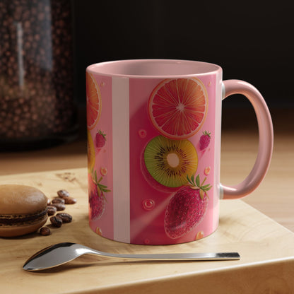A pink ceramic mug with a fruit pattern featuring strawberries, kiwi, and orange slices, displayed on a wooden surface next to a spoon and a macaron.