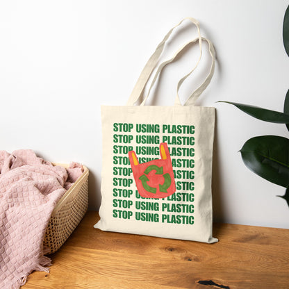 A natural canvas tote bag with green 'STOP USING PLASTIC' text and a red recycling symbol, displayed on a wooden table next to a pink blanket and a plant.