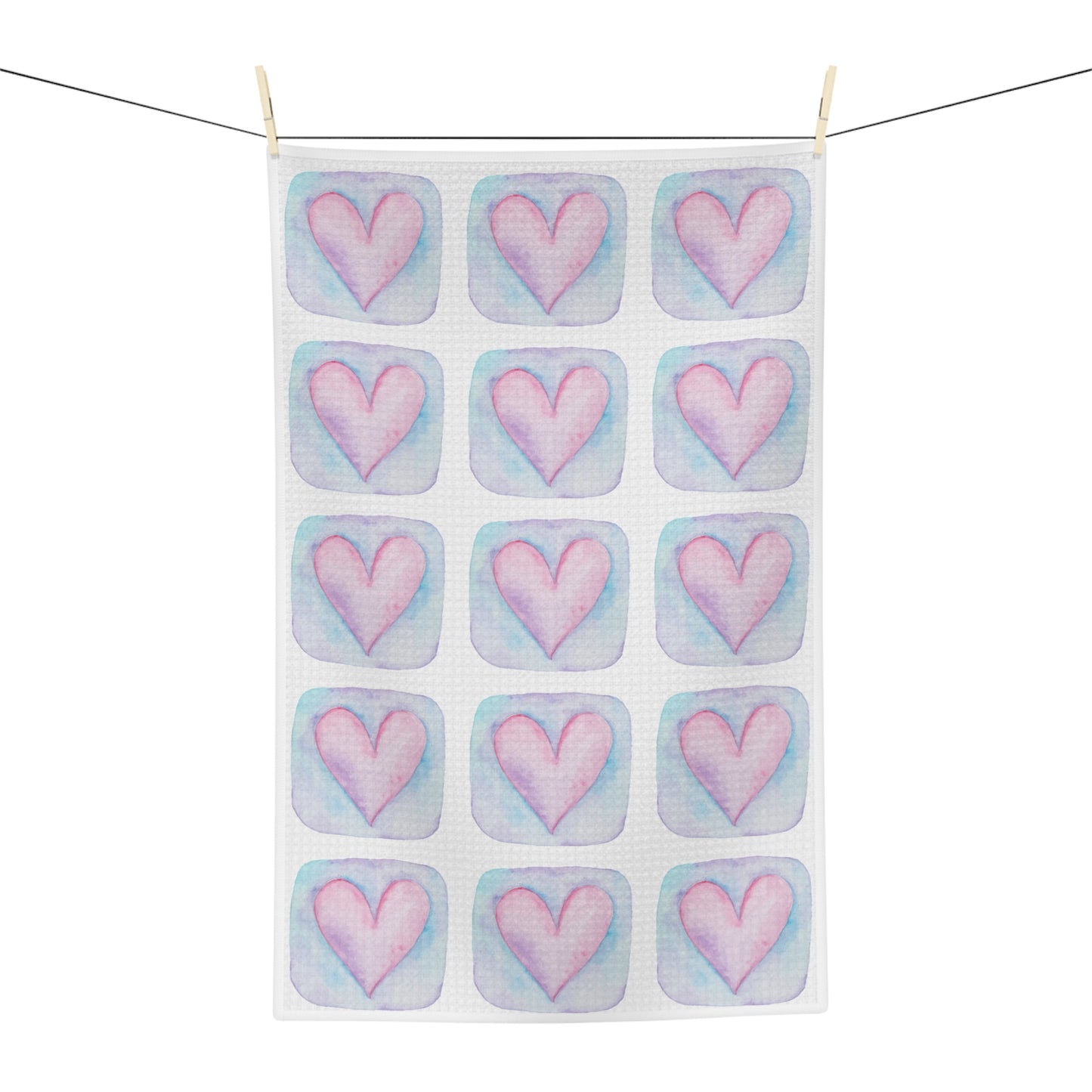 A white kitchen tea towel with a repeating pattern of pink hearts in pastel blue watercolor-style squares, hanging from a black clothesline with two wooden clips against a white background.