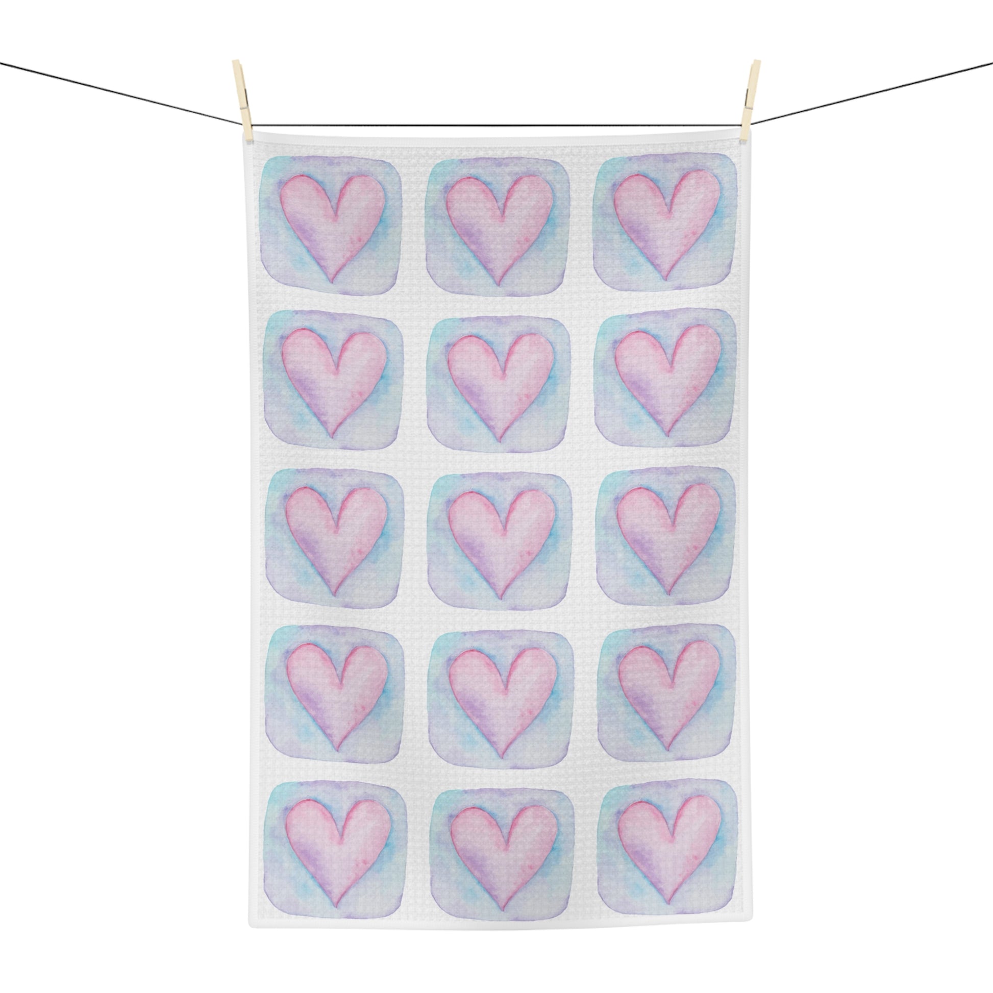 A white kitchen tea towel with a repeating pattern of pink hearts in pastel blue watercolor-style squares, hanging from a black clothesline with two wooden clips against a white background.