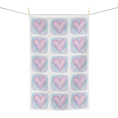 A white kitchen tea towel with a repeating pattern of pink hearts in pastel blue watercolor-style squares, hanging from a black clothesline with two wooden clips against a white background.