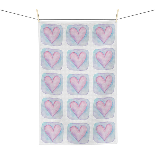 A white kitchen tea towel with a repeating pattern of pink hearts in pastel blue watercolor-style squares, hanging from a black clothesline with two wooden clips against a white background.