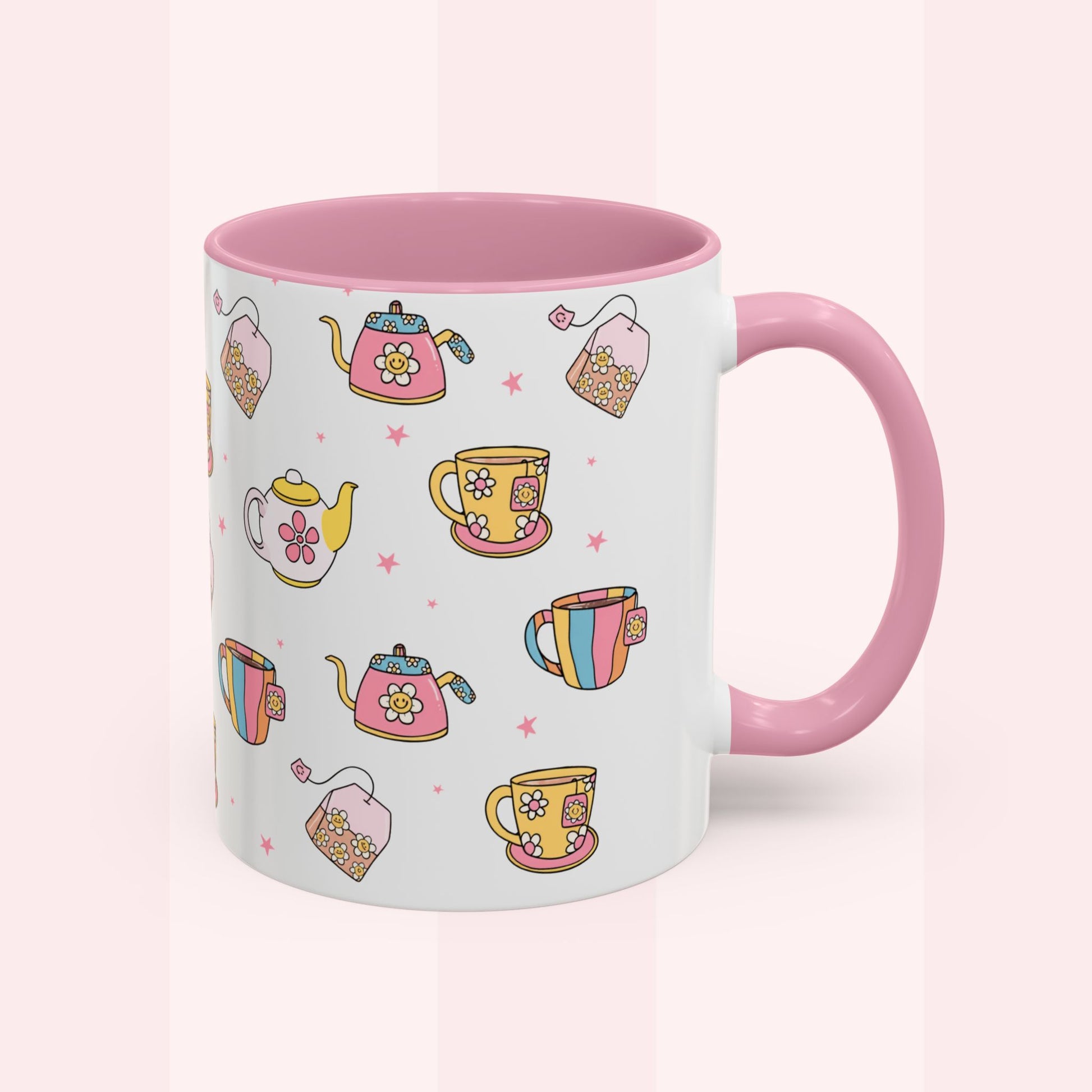A white ceramic mug with a pink interior and handle, decorated with a colorful pattern of teapots, teacups, and tea bags on a soft pink background.