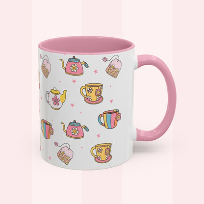 A white ceramic mug with a pink interior and handle, decorated with a colorful pattern of teapots, teacups, and tea bags on a soft pink background.