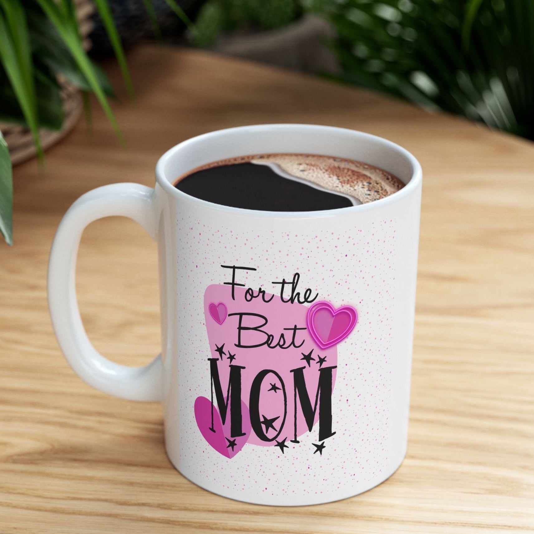 A white speckled ceramic mug filled with a dark coffee sits on a light wood table, decorated with the text 'For the Best MOM' in black and pink hearts. The background features a warm wooden surface and blurred green foliage.