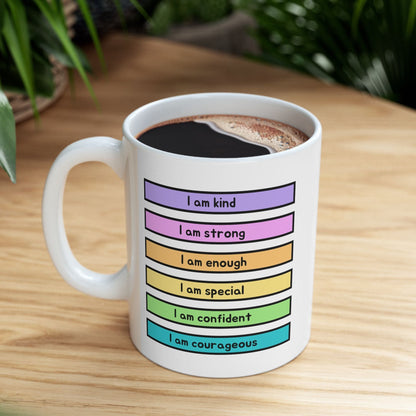 A white ceramic coffee mug filled with black coffee sits on a light wood table. The mug features a colorful graphic with horizontal rainbow-colored bars and text reading 'I am kind', 'I am strong', 'I am enough', 'I am special', 'I am confident', and 'I am courageous'.