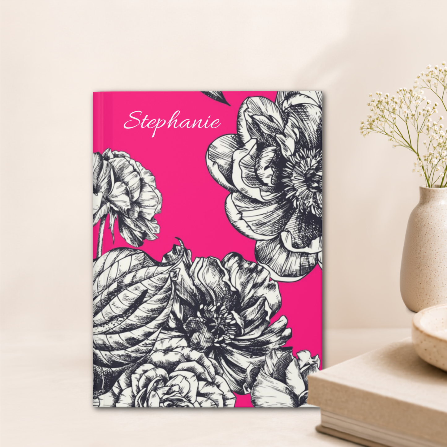 A personalized hardcover notebook with a bright pink cover, black and white floral illustration, and the name 'Stephanie' written in white script, styled on a tabletop next to a vase with dried flowers.