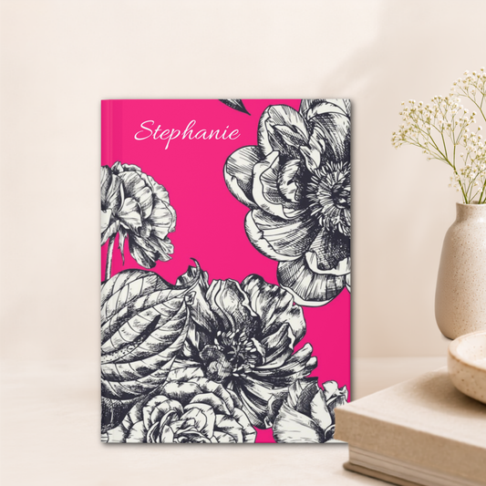 A personalized hardcover notebook with a bright pink cover, black and white floral illustration, and the name 'Stephanie' written in white script, styled on a tabletop next to a vase with dried flowers.
