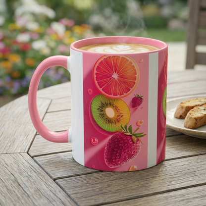 A pink ceramic coffee mug with a white vertical stripe and colorful fruit illustrations (grapefruit, kiwi, strawberry), filled with a hot latte on a wooden table with a blurred garden background.
