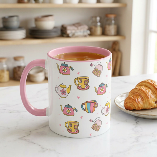 A white ceramic mug with a pink handle and interior, decorated with colorful teapot and teacup illustrations, filled with tea and placed on a marble countertop next to a croissant in a kitchen setting.
