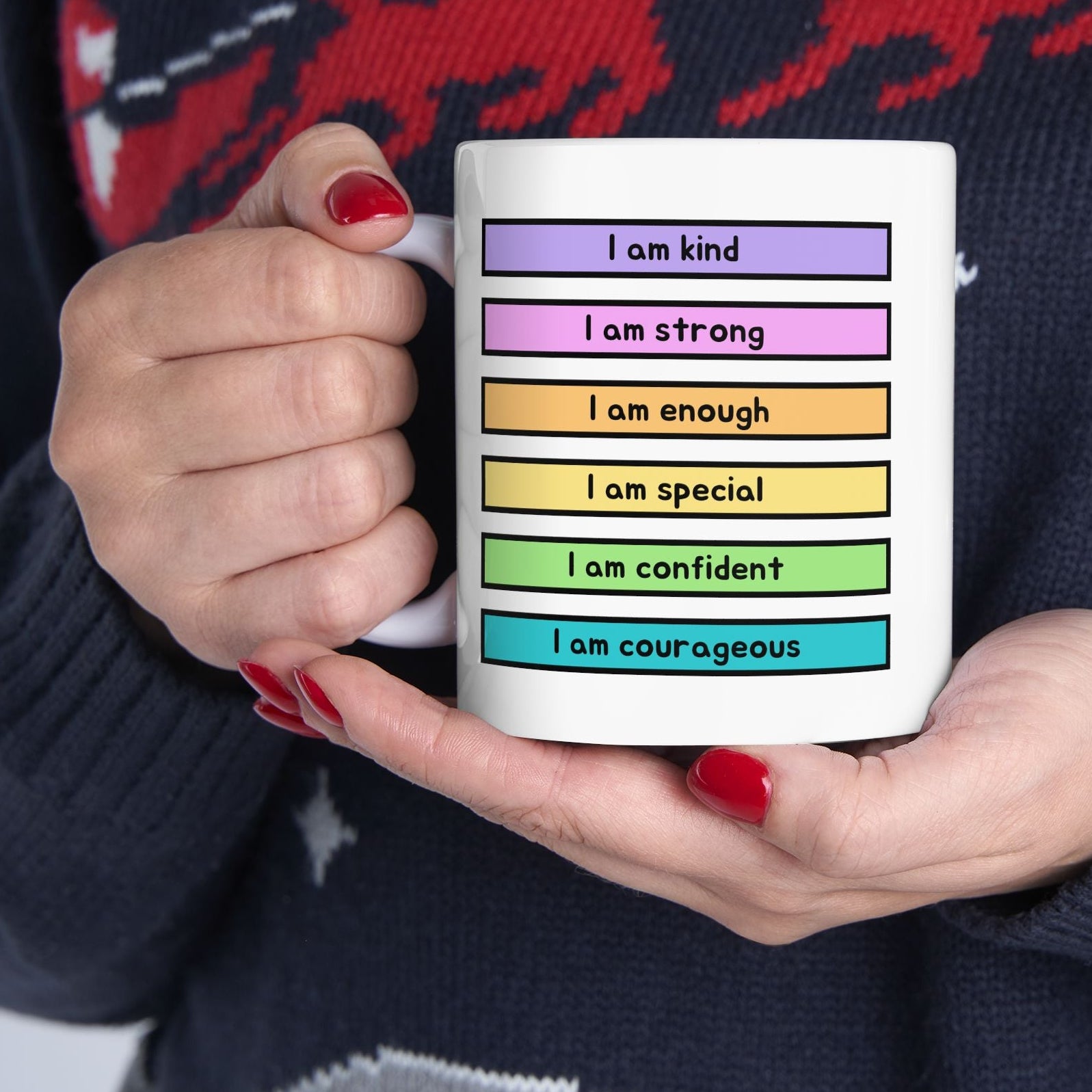 A person holding a white ceramic mug with colorful text blocks reading self-affirmations like 'I am kind', 'I am strong', 'I am enough', 'I am special', 'I am confident', and 'I am courageous'. The person is wearing a dark navy knit sweater with a red and gray holiday pattern.