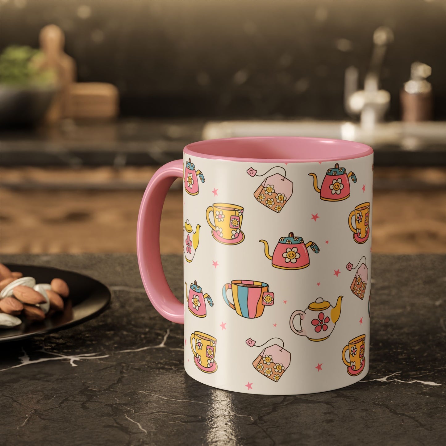 A white ceramic mug with a pink handle and pink interior, decorated with a colorful pattern of teapots, teacups, and tea bags on a dark kitchen countertop.