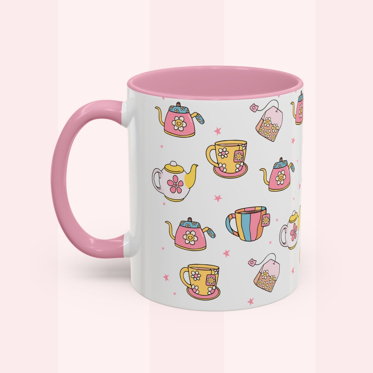 A white ceramic mug with a pink handle and pink interior, decorated with a colorful pattern of teapots, teacups, and tea bags on a pale pink background.