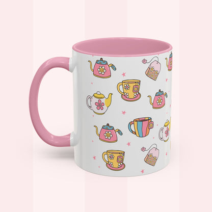 A white ceramic mug with a pink handle and pink interior, decorated with a colorful pattern of teapots, teacups, and tea bags on a pale pink background.