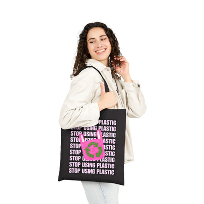 A woman wearing a light beige jacket and jeans holds a black tote bag with a pink and green graphic that reads 'STOP USING PLASTIC' in white text. The background is solid white.