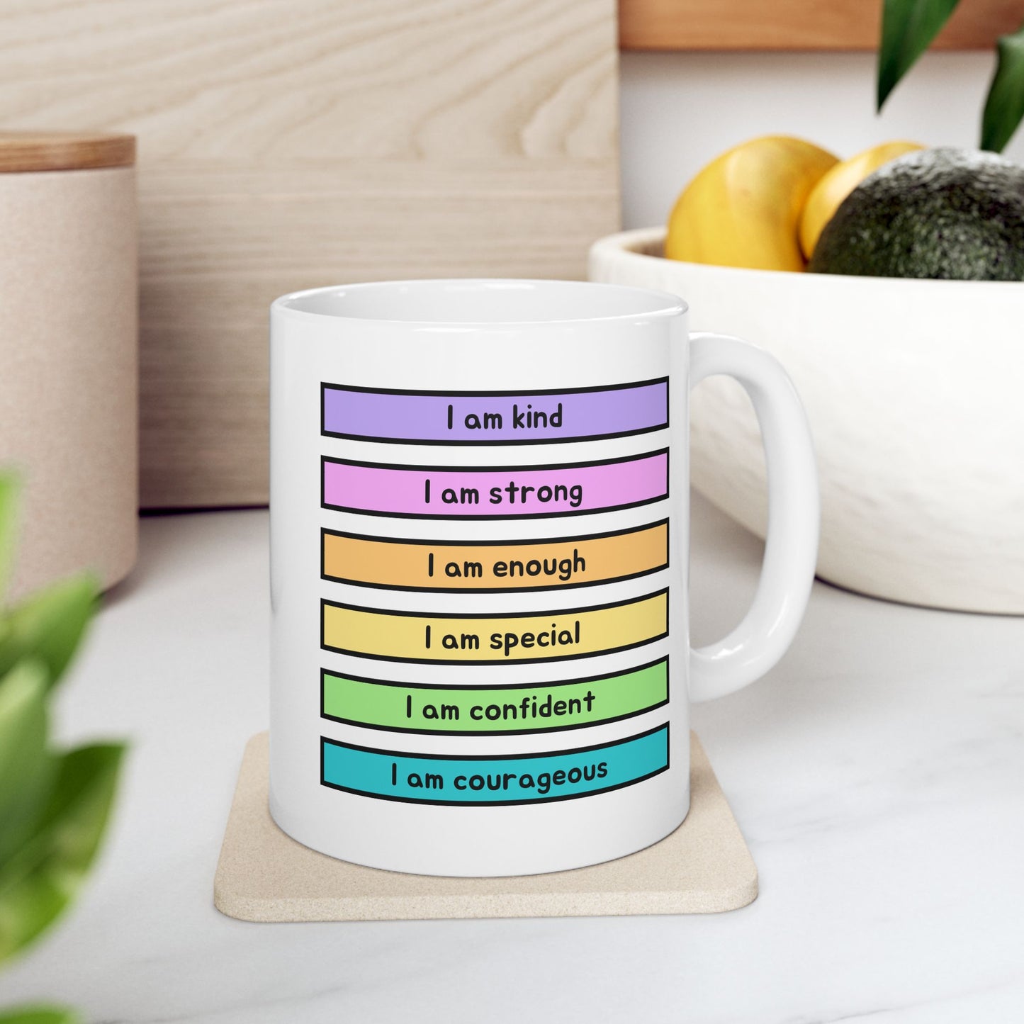 A white ceramic mug with colorful horizontal stripes and motivational text, sitting on a coaster on a kitchen counter with a bowl of fruit in the background.