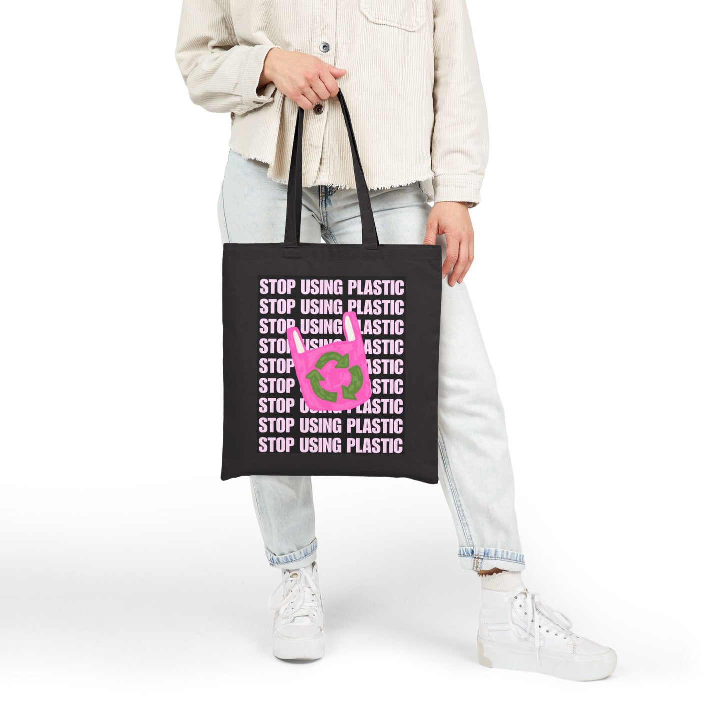 A person wearing a light beige jacket, light wash jeans, and white sneakers holds a black canvas tote bag with the text 'STOP USING PLASTIC' repeated in white and a pink plastic bag icon in the center, against a plain white background.