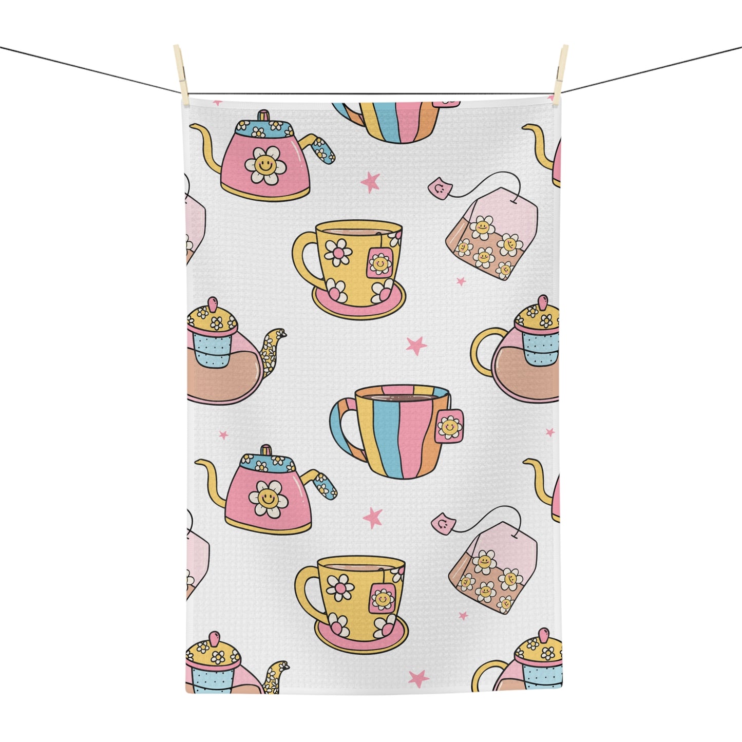 A rectangular tea towel with a waffle texture, featuring a colorful pattern of teapots, teacups, teabags, and pink stars on a white background, hanging from a clothesline with wooden clips.