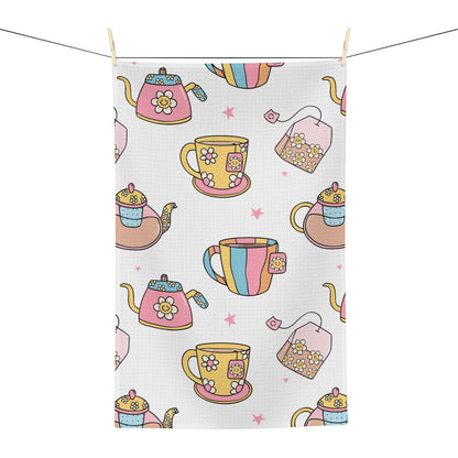 A rectangular tea towel with a waffle texture, featuring a colorful pattern of teapots, teacups, teabags, and pink stars on a white background, hanging from a clothesline with wooden clips.