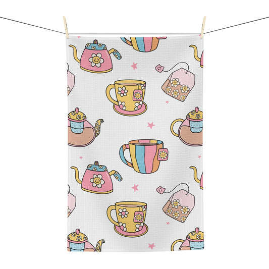 A rectangular tea towel with a waffle texture, featuring a colorful pattern of teapots, teacups, teabags, and pink stars on a white background, hanging from a clothesline with wooden clips.