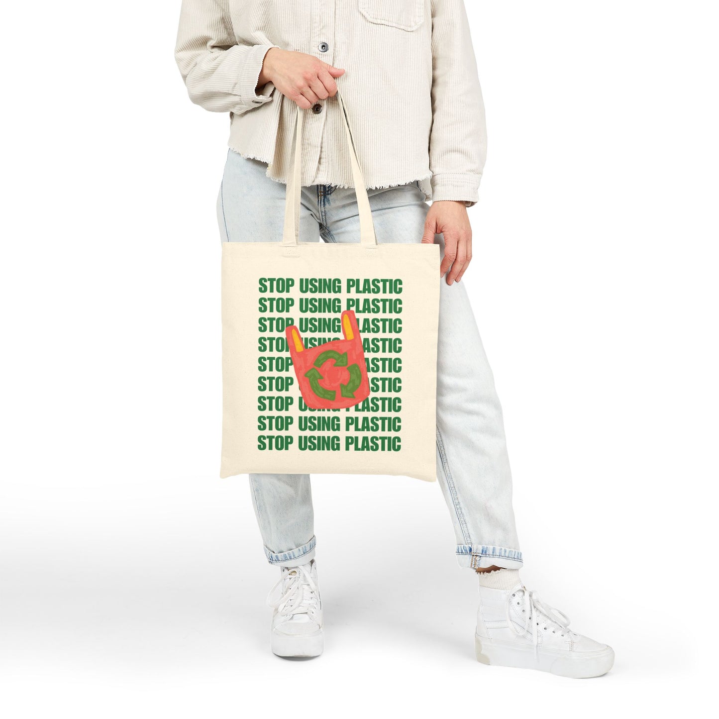 A person wearing a light beige jacket, light blue jeans, and white sneakers holds a cream canvas tote bag printed with the green text 'STOP USING PLASTIC' and a red-and-green recycling symbol on the front, against a plain white studio background.
