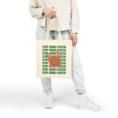 A person wearing a light beige jacket, light blue jeans, and white sneakers holds a cream canvas tote bag printed with the green text 'STOP USING PLASTIC' and a red-and-green recycling symbol on the front, against a plain white studio background.