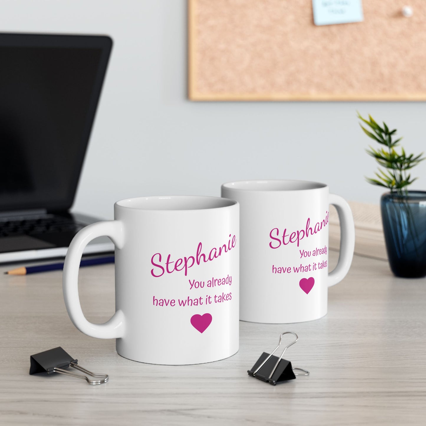 Two white mugs with pink text on a desk with a laptop and plant in the background.