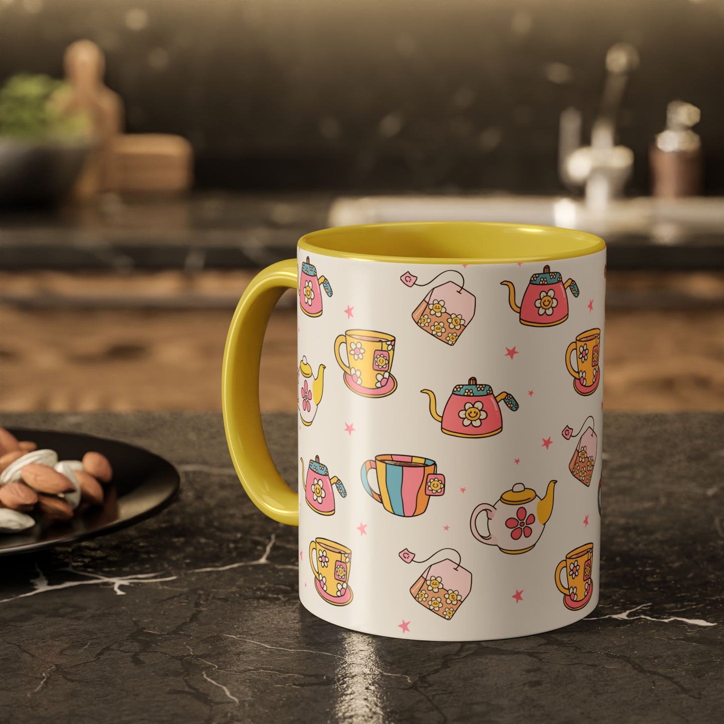 A white ceramic mug with a yellow handle and yellow interior, decorated with a colorful pattern of teapots, teacups, and tea bags, sitting on a dark kitchen countertop.