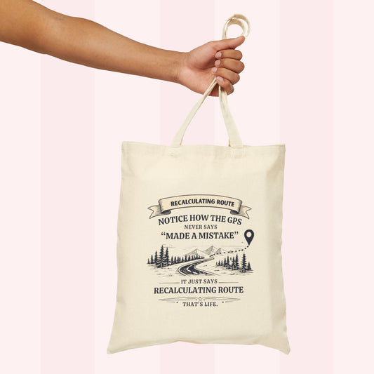 An off-white canvas tote bag with a black graphic print and text, held by a person's hand against a light pink striped background.