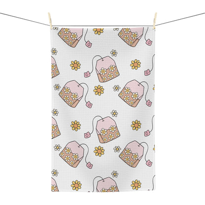 A white kitchen tea towel with a repeating pattern of pink teabags filled with yellow and white flowers, hanging from a string with wooden clips against a white background.