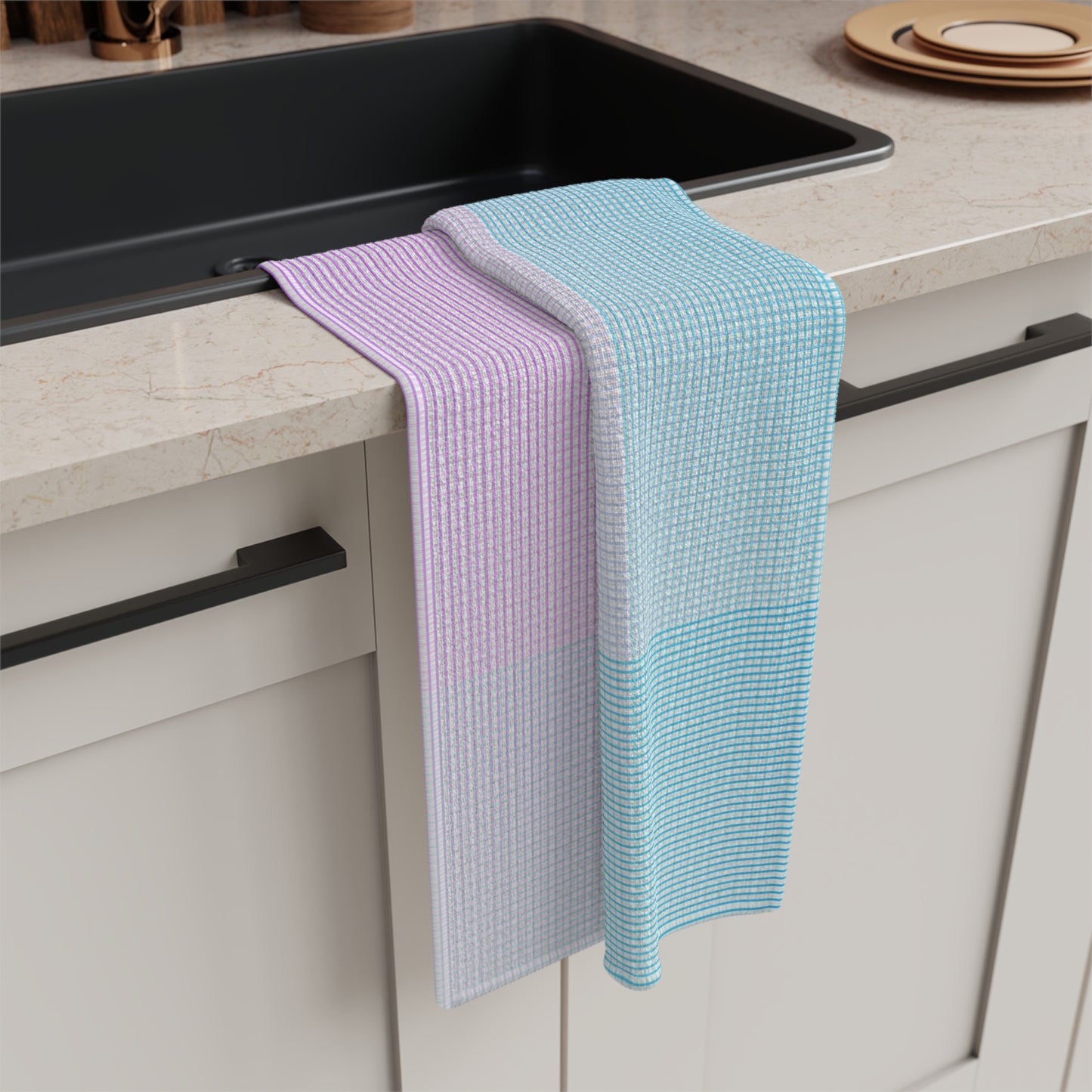 Two waffle-weave kitchen towels draped over the edge of a black sink in a modern kitchen setting. One towel is lavender and white, and the other is light blue and white.
