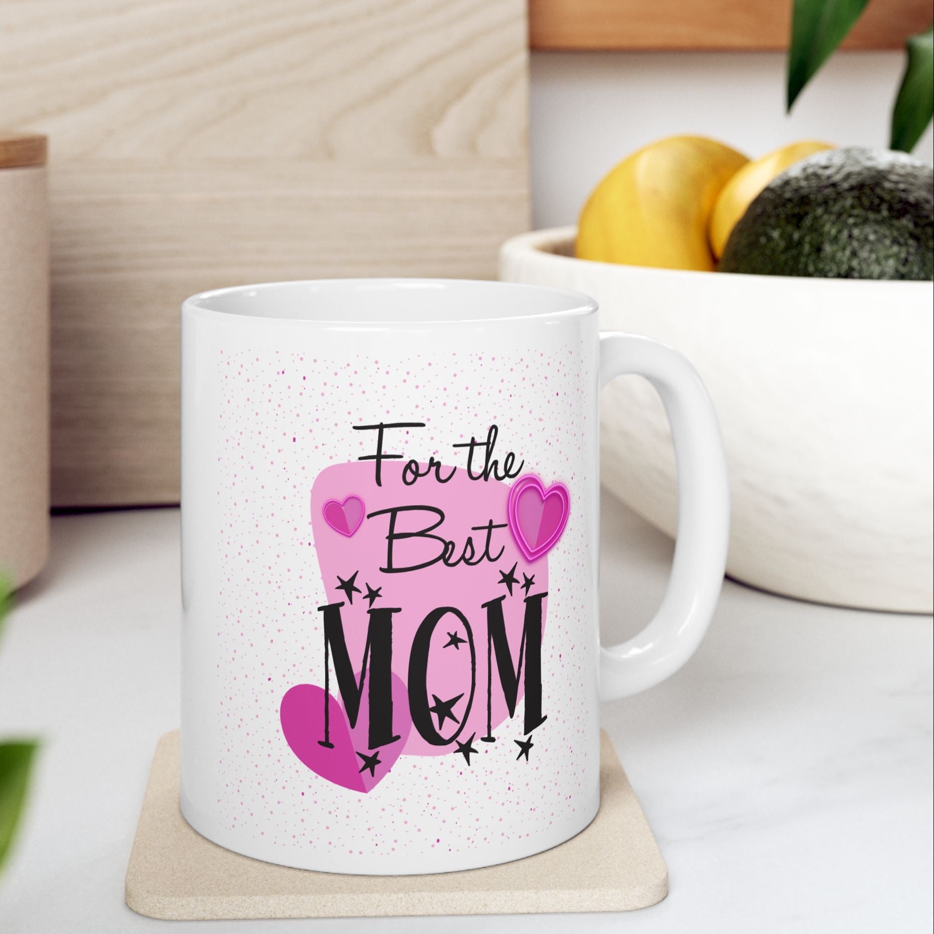 A white ceramic mug with a pink and black 'For the Best MOM' graphic, resting on a light wood coaster on a kitchen counter with blurred background elements.