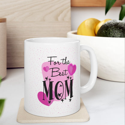 A white ceramic mug with a pink and black 'For the Best MOM' graphic, resting on a light wood coaster on a kitchen counter with blurred background elements.