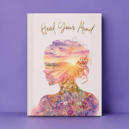 A hardcover journal or notebook standing upright against a lavender-purple background. The cover is white with the phrase 'Heal Your Mind' in gold script at the top and a large watercolor-style silhouette of a woman's profile filled with a colorful sunset and floral design.