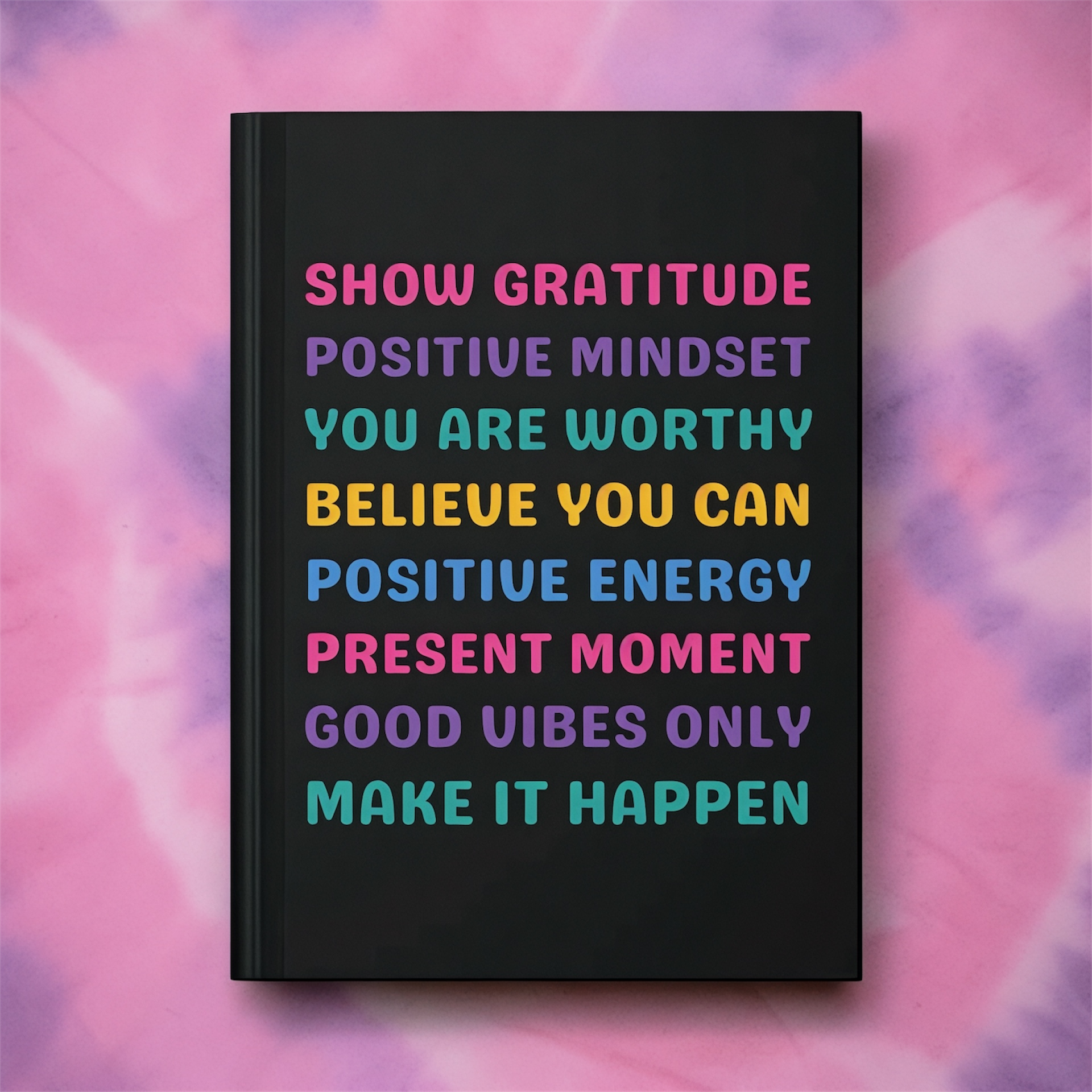 A black hardcover notebook or journal with colorful motivational text on the cover, displayed against a pink and purple tie-dye background.