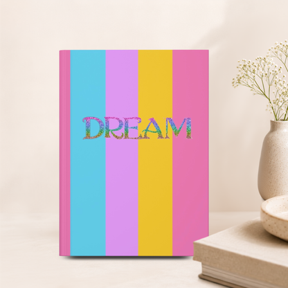 A hardcover notebook with vertical pastel stripes in blue, purple, yellow, and pink, featuring the word 'DREAM' in glittery multicolor letters across the center. The notebook is styled on a light beige surface next to a small vase with dried flowers and a book.