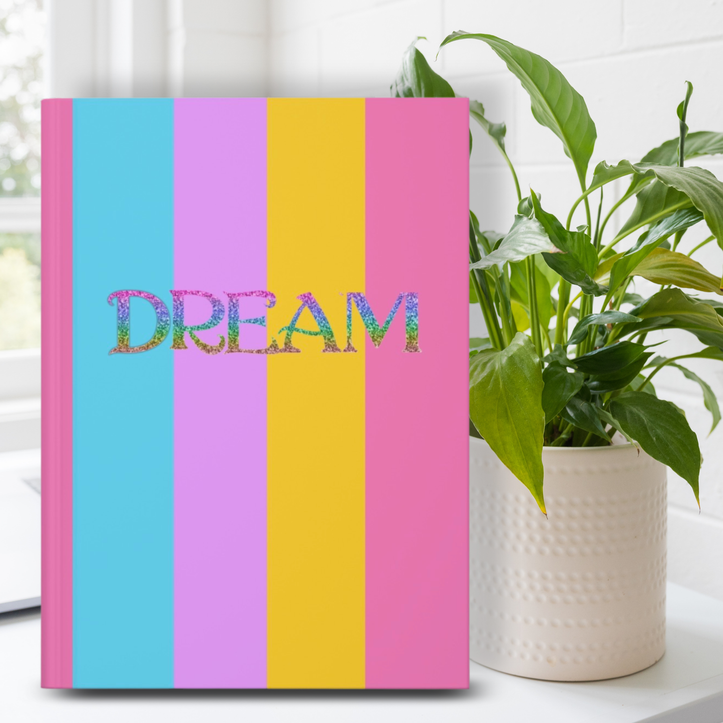 A hardcover notebook or journal with vertical pastel stripes in pink, blue, lavender, and yellow, featuring the word 'DREAM' in glittery multicolor letters across the front. It is styled on a white surface next to a potted green plant.