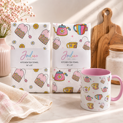Two Jodu Designs kitchen tea towels with a colorful pattern of teabags, teapots, and flowers are shown next to a matching pink-handled mug on a light beige surface.